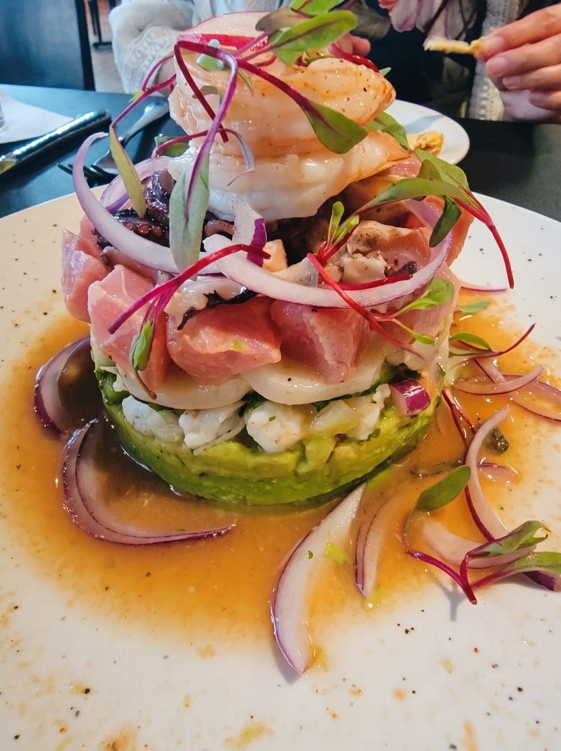 Stacked seafood ceviche with shrimp, tuna, avocado, red onion, and microgreens on a white plate.