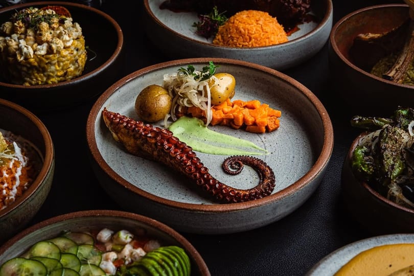 Grilled octopus with potatoes, sweet potato, and green sauce, surrounded by other dishes.