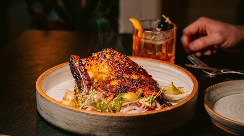 Steaming roasted chicken with vegetables and a cocktail, with a hand reaching for a fork.