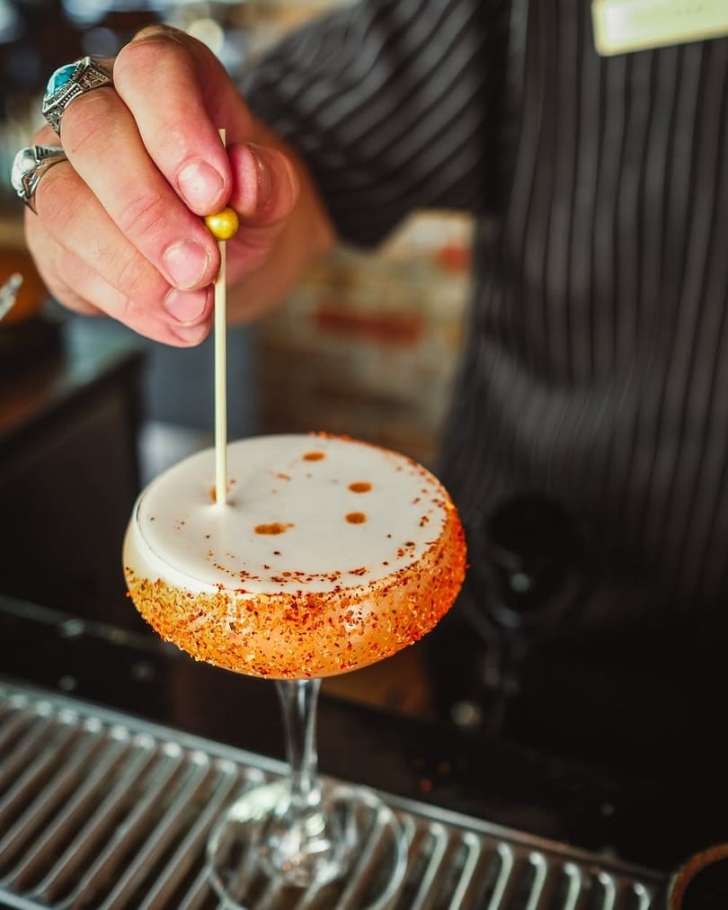 Bartender garnishing a cocktail with white foam, chili-dusted rim, and a golden bead on a pick.