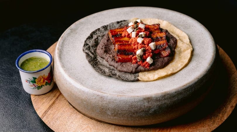 Pork belly with red sauce, black beans, and cheese on a tortilla, with green salsa.