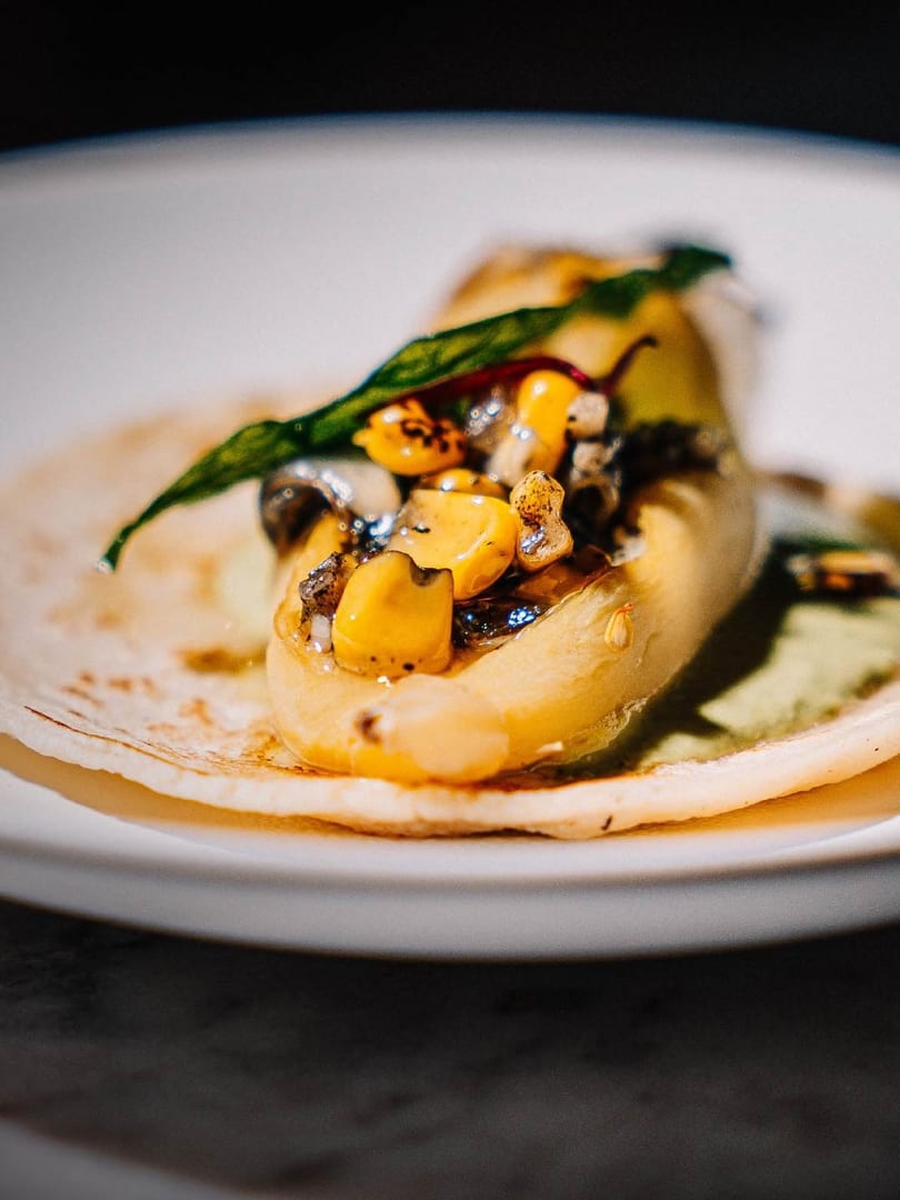 Close-up of a taco with corn, mushrooms, and a green leaf on a white plate.