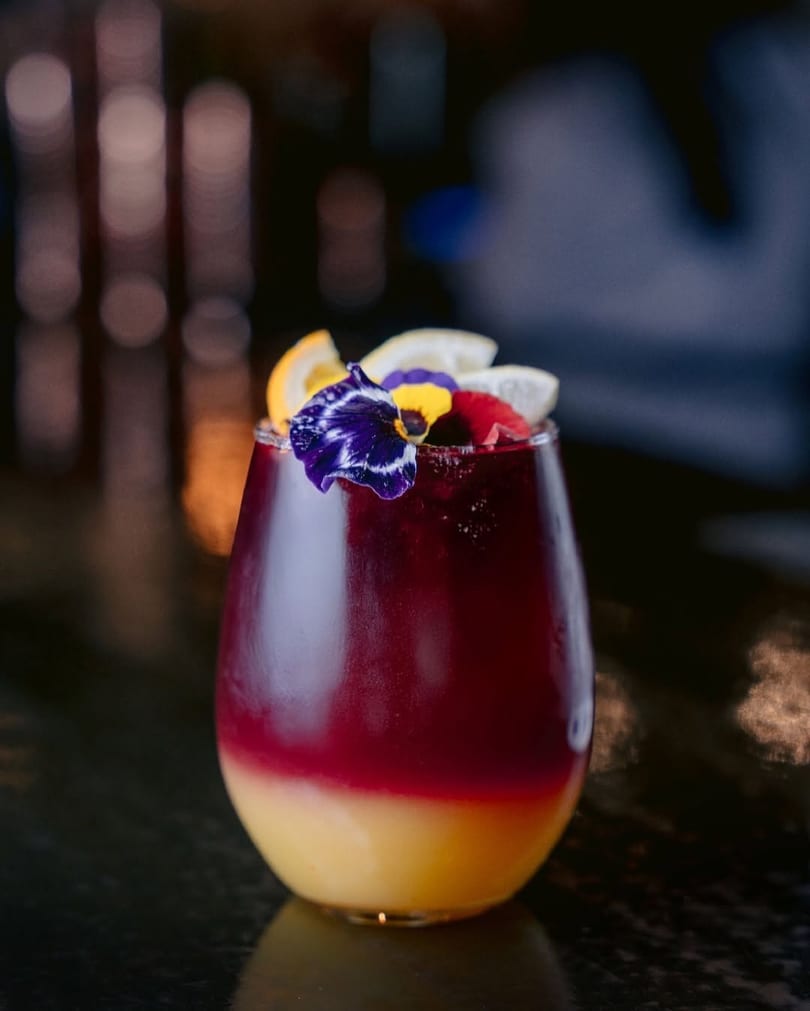Layered red and yellow cocktail garnished with citrus slices and edible flowers on a dark surface.