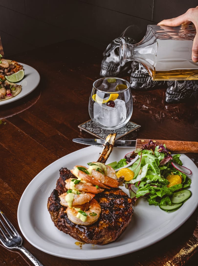Grilled steak with shrimp and salad, a cocktail, and tacos on a dark wooden table.