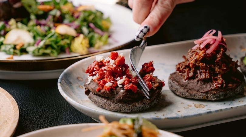 A hand with painted nails uses a fork to cut into a red and white topped appetizer.